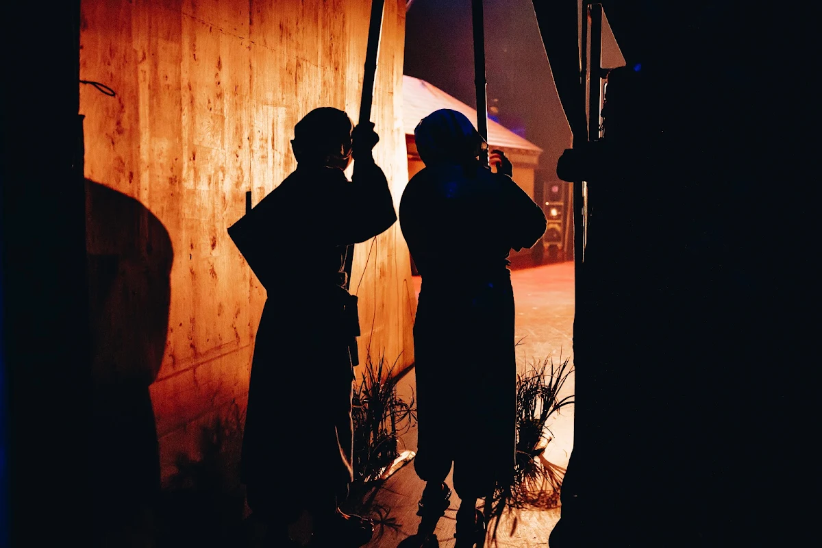 Two hooded figures silhouetted against a warmly lit wooden wall of the set