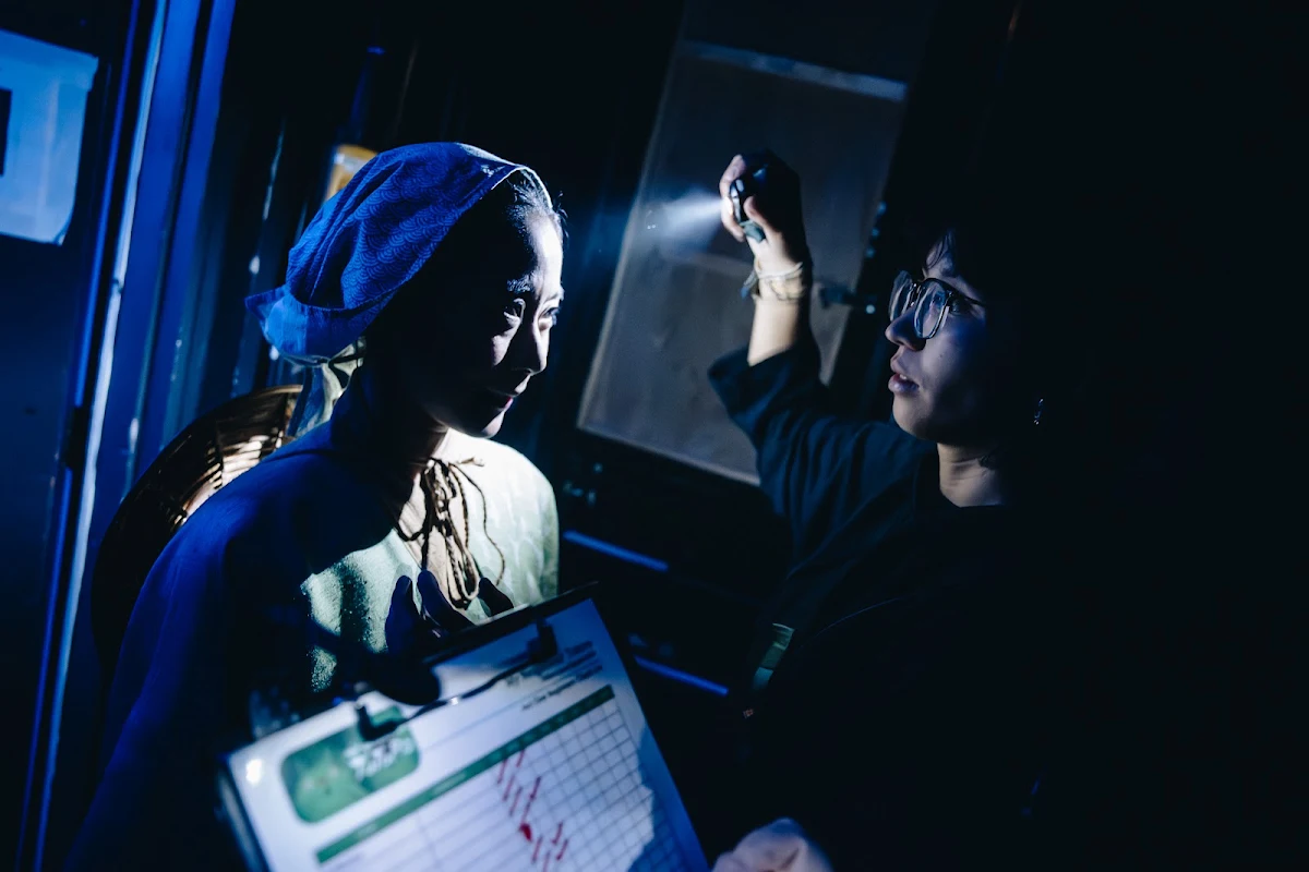 Natsumi Kuroda in a headscarf lit by blue light in the wings, as a crew member with a torch checks a clipboard