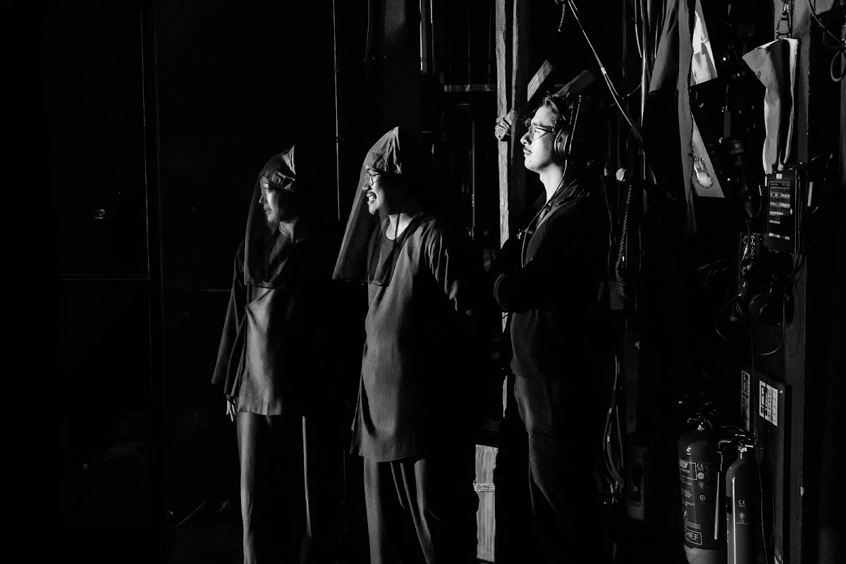 Black and white photograph of hooded performers standing in the dark of the wings
