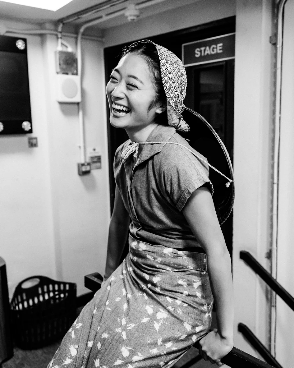 Black and white photograph of Sera Maehara in a headscarf and patterned dress laughing beside a door marked Stage