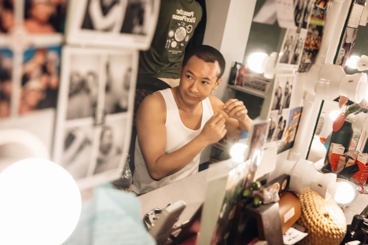 Steven Nguyen in a white vest getting ready at a dressing room mirror covered in photographs