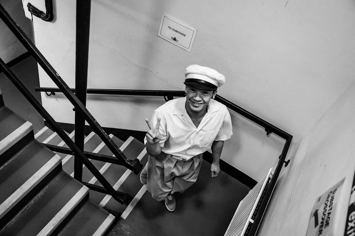 Black and white photograph looking down a stairwell at Steven Nguyen in a white cap, making a peace sign