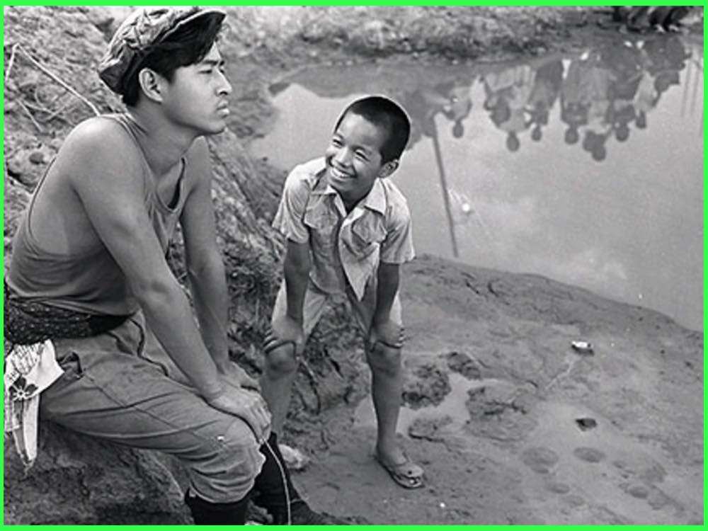 Black and white still from My Second Brother (1959): a young man in a vest sits on a muddy bank as a smiling boy leans towards him