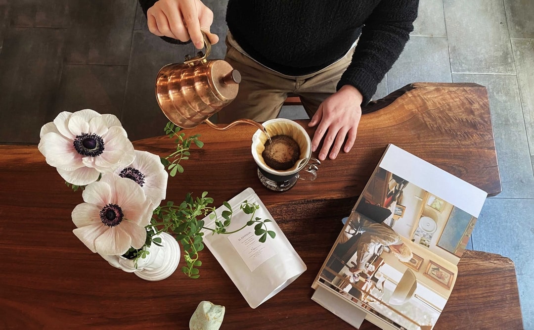 A barista pouring a hand drip coffee from a copper kettle on a wooden table with white anemones at Nem Coffee and Espresso