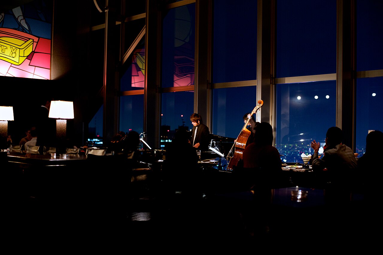 A jazz pianist and double bass player performing in the dimly lit New York Bar at Park Hyatt Tokyo, with the city lights beyond its tall windows