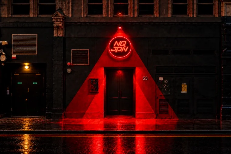 The black frontage of NQJPN at 53 Berwick Street in Soho at night, with a red neon NQJPN sign above the door and a red triangle of light reflected on the wet pavement