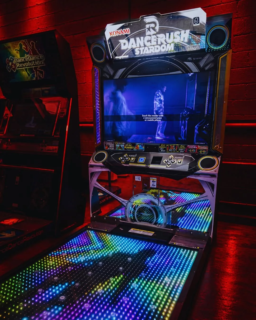 A Konami DANCERUSH STARDOM cabinet with a tall screen above a large dance platform lit in rainbow LED colours