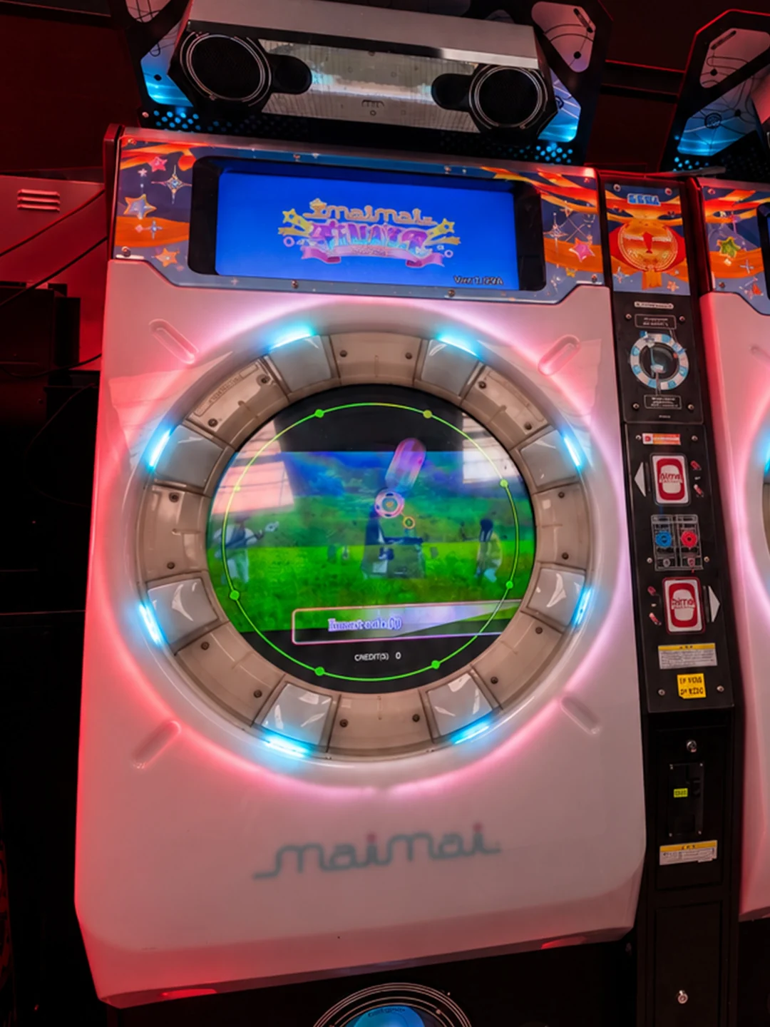 A white Sega maimai cabinet with a round touchscreen ringed by glowing blue buttons, lit by red light inside NQJPN's arcade