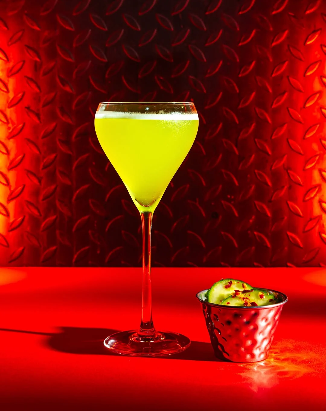 A pale green Melon and Cucumber Daiquiri in a stemmed coupe glass beside a small metal cup of melon balls, against a red chequer plate wall