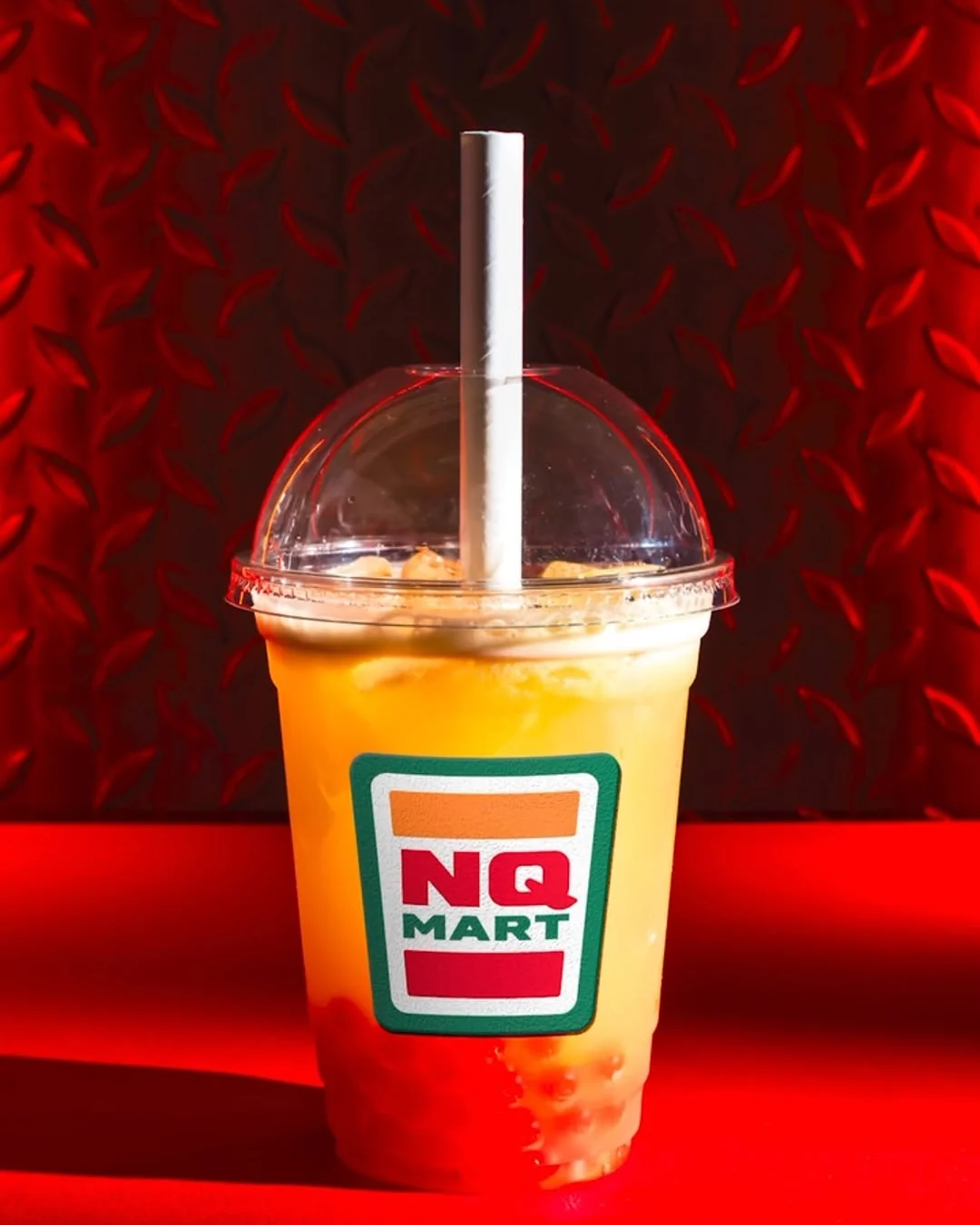 An orange Tequila, Apple and Yuzu Bubble Tea in a domed plastic cup with a green, red and orange NQ Mart label, against a red wall