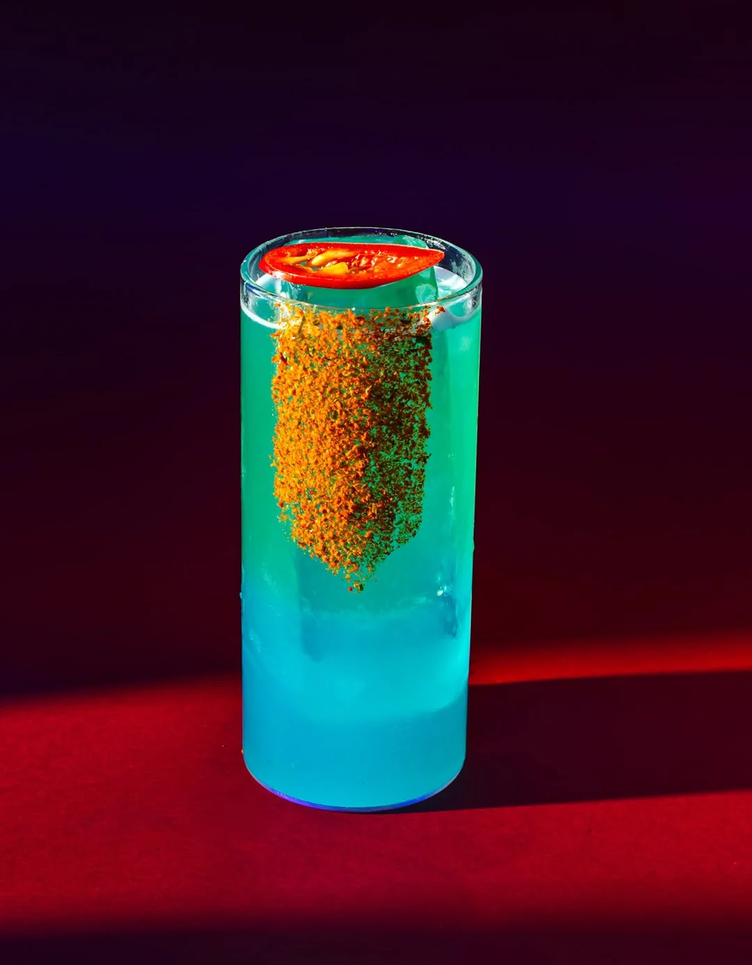 A tall bright blue Shichimi Hi-Ball with a stripe of orange seven-spice seasoning running down the outside of the glass, on a red surface