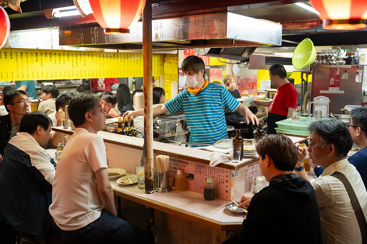 Diners sitting shoulder to shoulder at a small counter in Omoide Yokocho, Shinjuku, while the cook works the grill