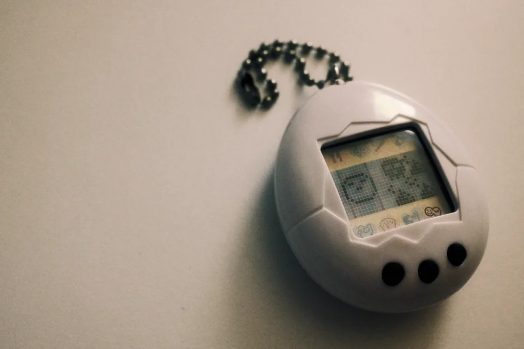 White egg shaped Tamagotchi Original on a ball chain, its pixel screen lit, against a pale grey background