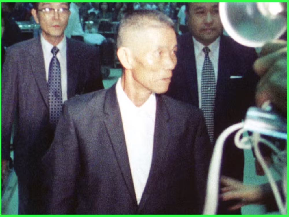 Colour still from Outlaw-Matsu Returns Home (1973): a thin man in a dark suit walks through a crowd of men in suits and press photographers