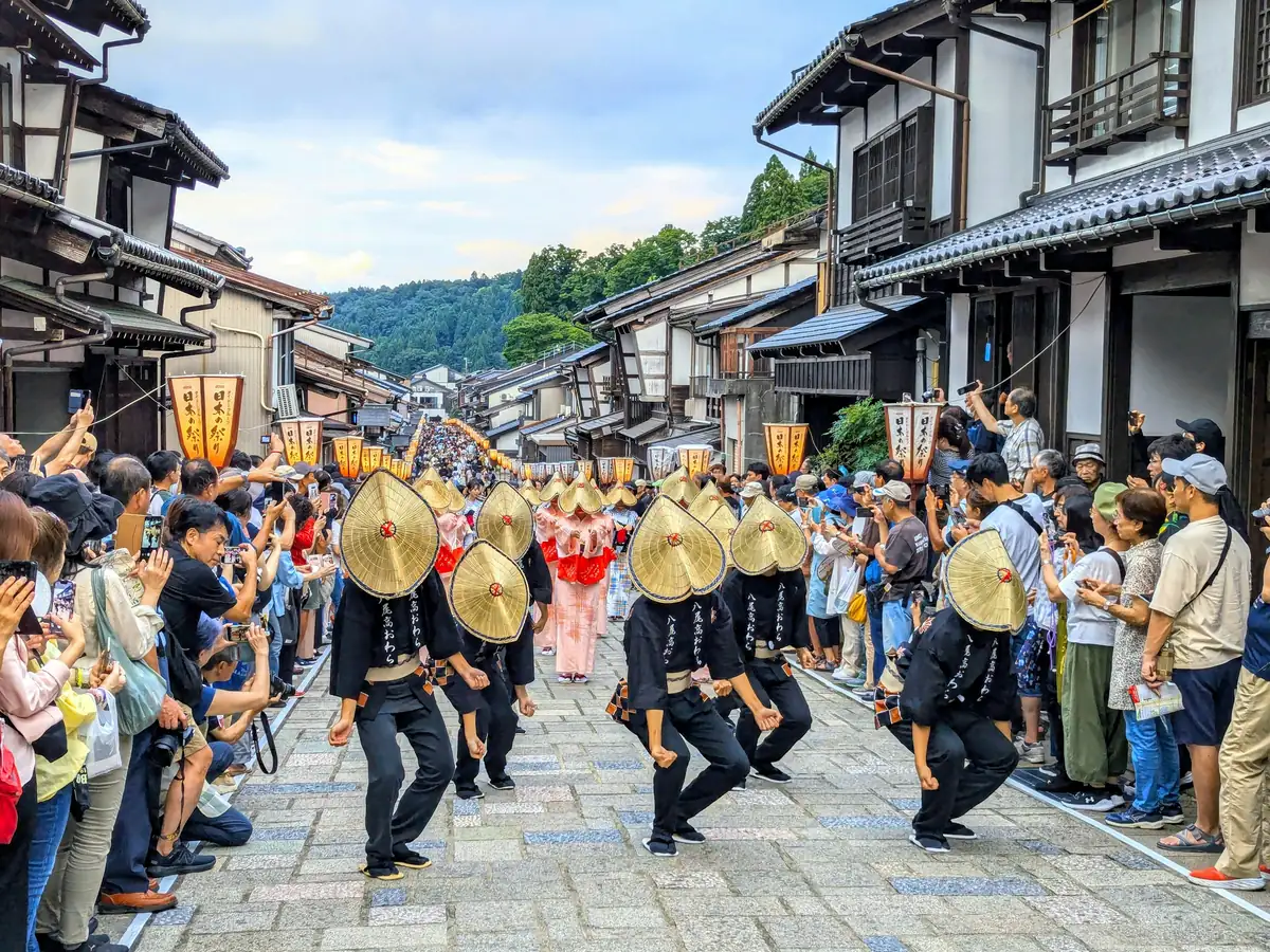 Dancers in straw hats and dark yukata moving in a line down a stone street lined with wooden townhouses and paper lanterns during Owara Kaze no Bon in Yatsuo, Toyama, watched by crowds on both sides