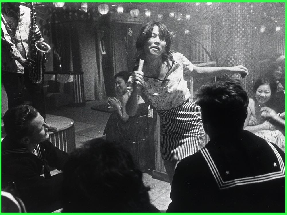 Black and white still from Pigs and Battleships (1961): a young woman dances in a crowded bar as sailors watch