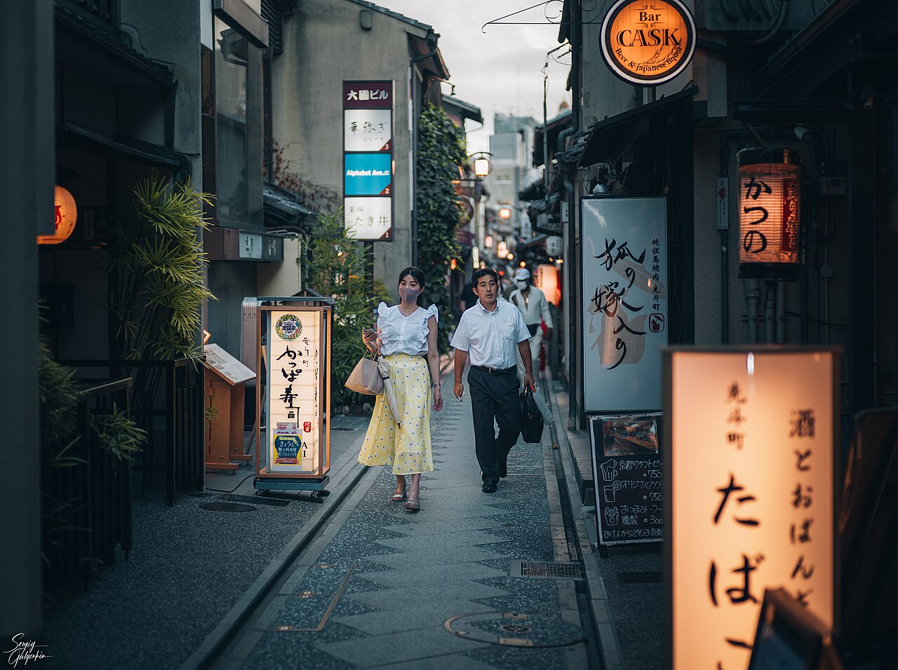 People walking along the narrow stone-paved Pontocho alley in Kyoto, lined with lit restaurant and bar signs