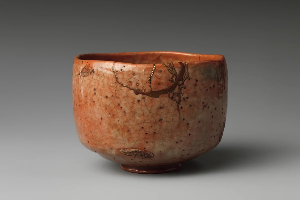 A red Raku tea bowl with a speckled orange glaze and straight, slightly uneven walls, a crack across its side mended with a thin line of gold