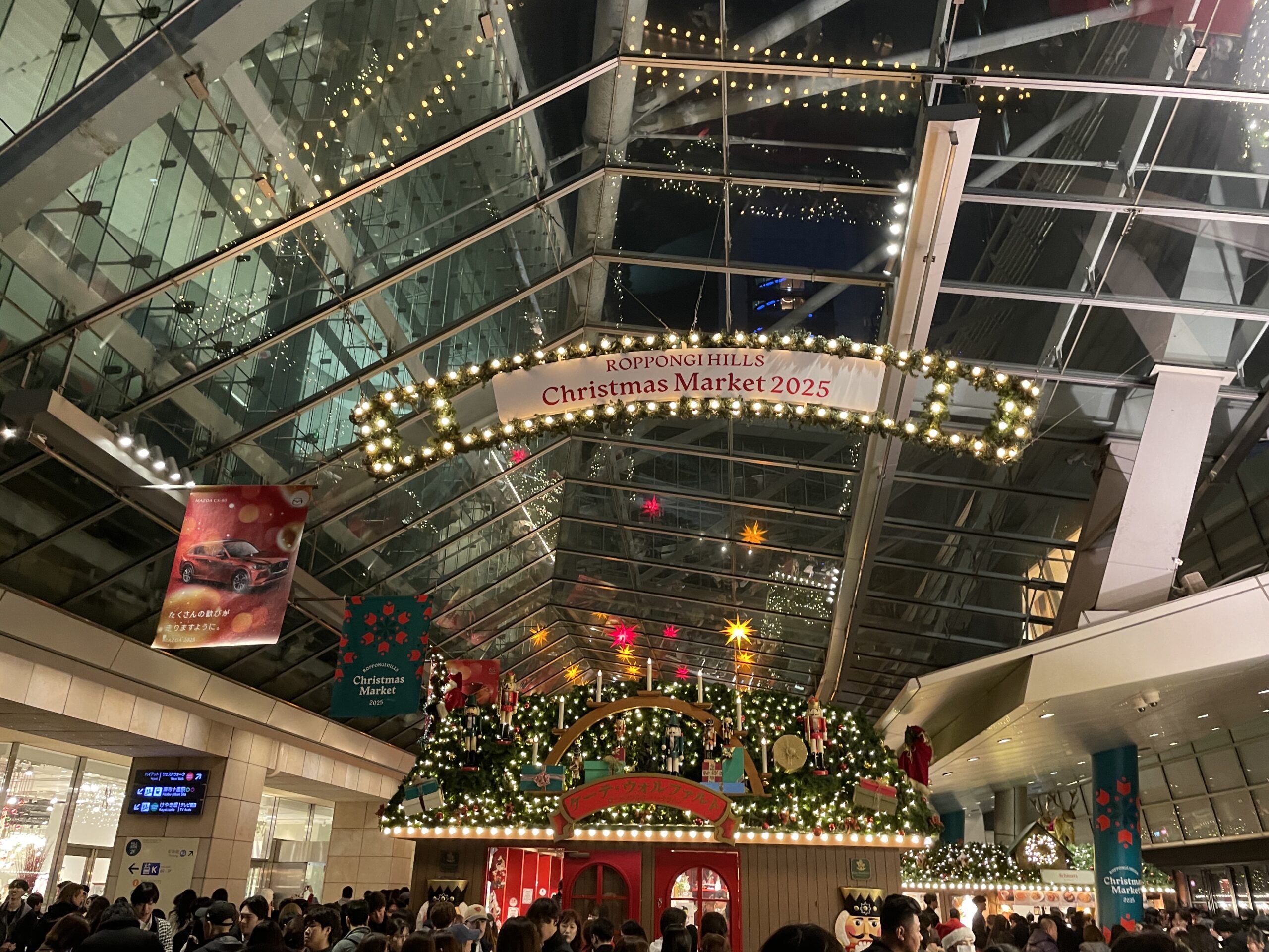 The Roppongi Hills Christmas Market 2025 sign hanging under the glass roof of Oyane Plaza in Tokyo, with decorated wooden stalls and crowds below