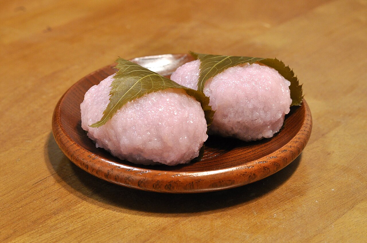 Two pink sakura mochi rice cakes wrapped in pickled cherry leaves on a wooden plate