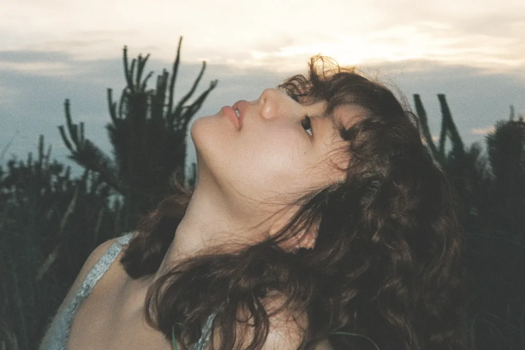 Salasa tilting her head back to look up at an evening sky, with wavy brown hair and dark plants behind her