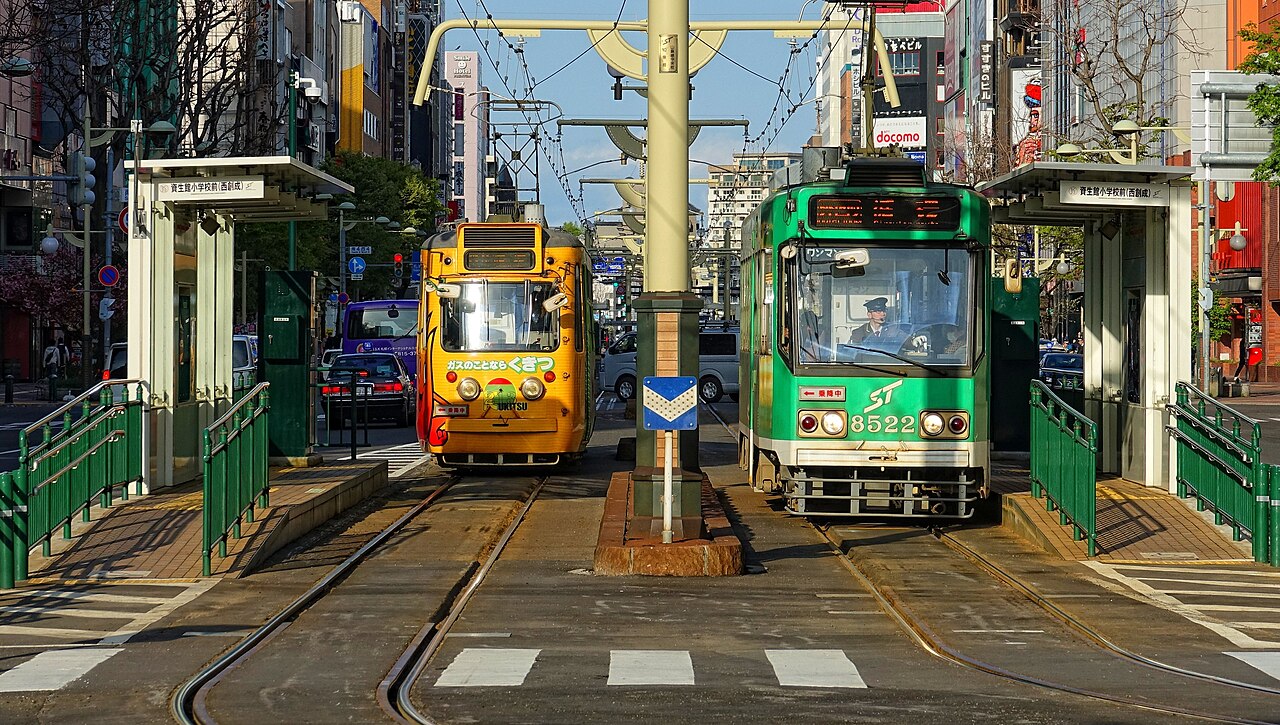 Two Sapporo streetcars, one yellow and one green, at a tram stop in the middle of a downtown Sapporo street