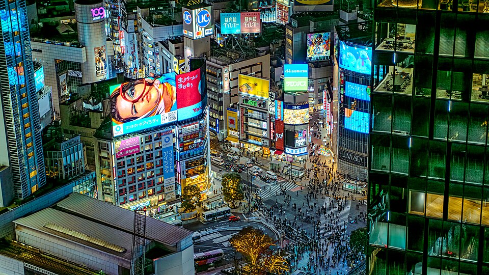 Shibuya Crossing in Tokyo seen from above at night, with crowds crossing beneath bright advertising screens