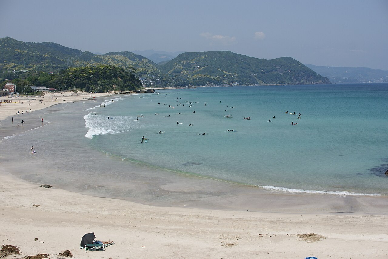 Surfers and swimmers in the clear water of the curving bay at Shirahama Ohama Beach in Shimoda, with green hills behind