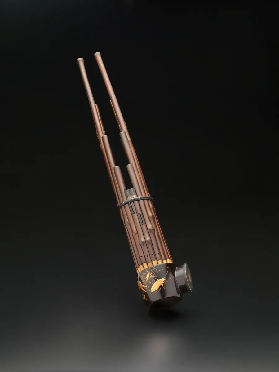A shō mouth organ, a cluster of slim bamboo pipes rising from a lacquered wind chest, photographed against black.