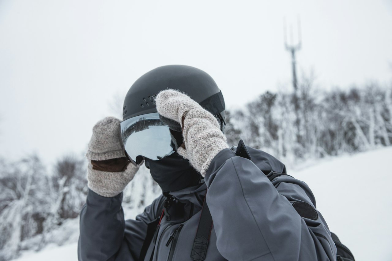 A skier in a helmet, face cover and wool mittens adjusting mirrored goggles on a snowy slope