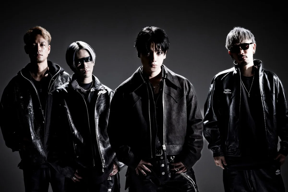 The four members of Japanese rock band SPYAIR standing side by side in black leather jackets against a dark grey studio backdrop, one wearing sunglasses and one with a black band painted across his eyes