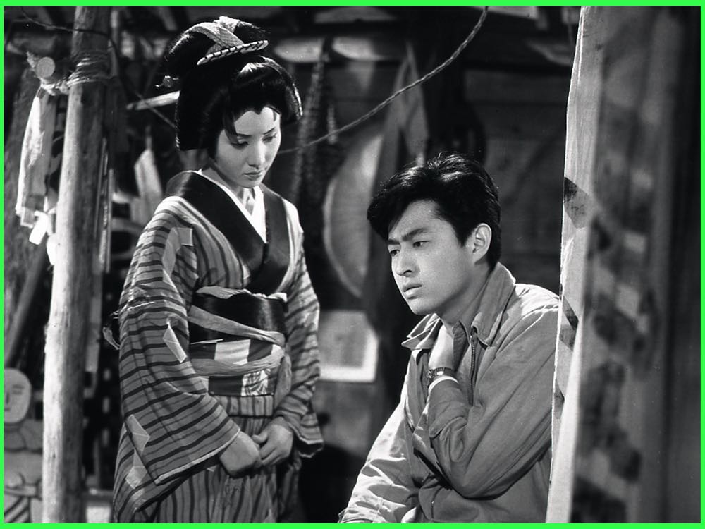 Black and white still from Stolen Desire (1958): a young woman in a striped kimono looks down beside a young man in a work shirt at a travelling theatre