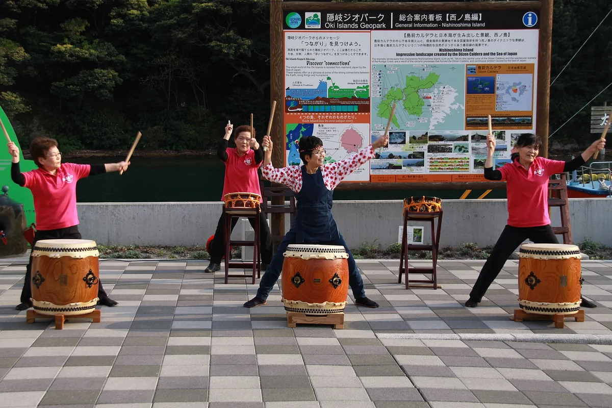 Four women in red and pink shirts strike barrel-shaped taiko drums on low stands on a paved harbour front, arms raised with sticks in the air, in front of an Oki Islands Geopark information board