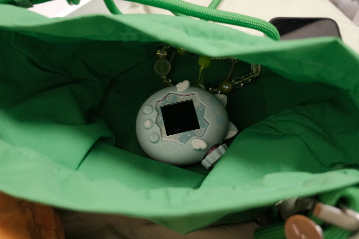Pale blue Tamagotchi on a beaded chain inside an open green bag