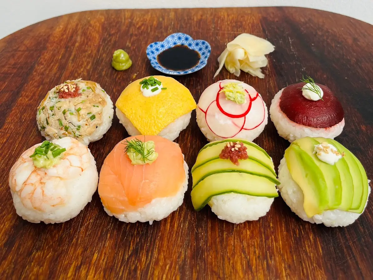 Eight balls of temari sushi on a wooden board, topped with salmon, prawn, avocado, cucumber and egg, with pickled ginger, wasabi and a dish of soy sauce
