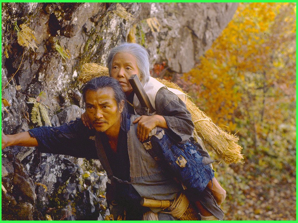 Colour still from The Ballad of Narayama (1983): a man carries his elderly mother on his back up a rocky mountainside in autumn