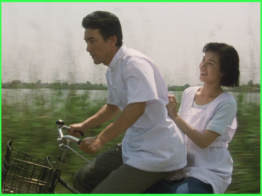 Colour still from The Eel (1997): Kōji Yakusho rides a bicycle with Misa Shimizu on the back, a river behind them