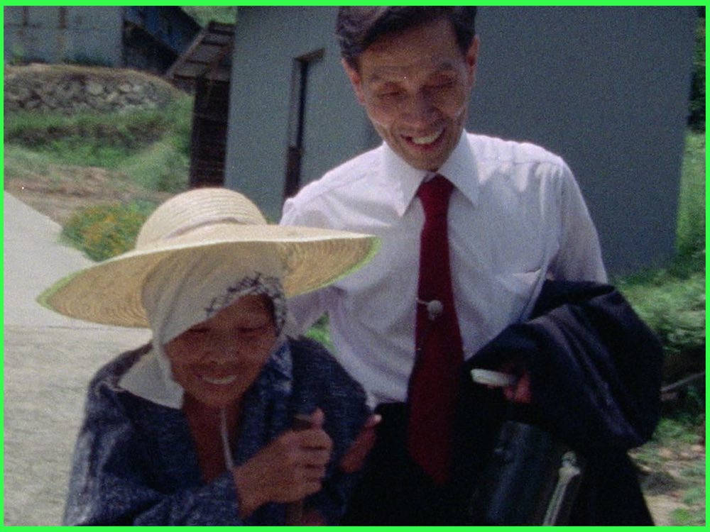 Colour still from The Emperor's Naked Army Marches On (1987): a smiling man in a white shirt and red tie walks beside an elderly woman in a straw hat