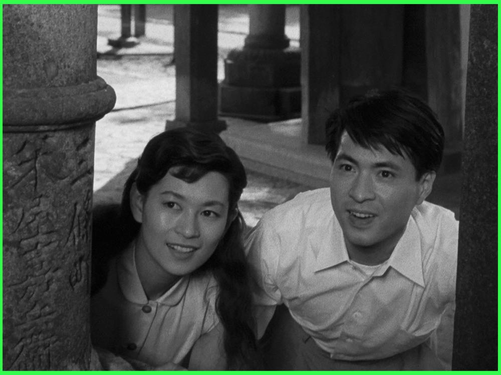 Black and white still from The Moon Has Risen (1955): a young woman and a young man peer out from behind a stone pillar