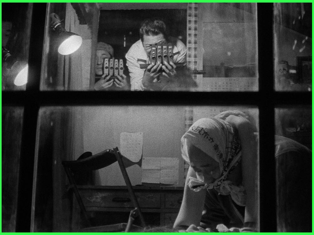 Black and white still from The Pornographers (1966): seen through a window, a man holds up reels of film while a woman in a headscarf works below