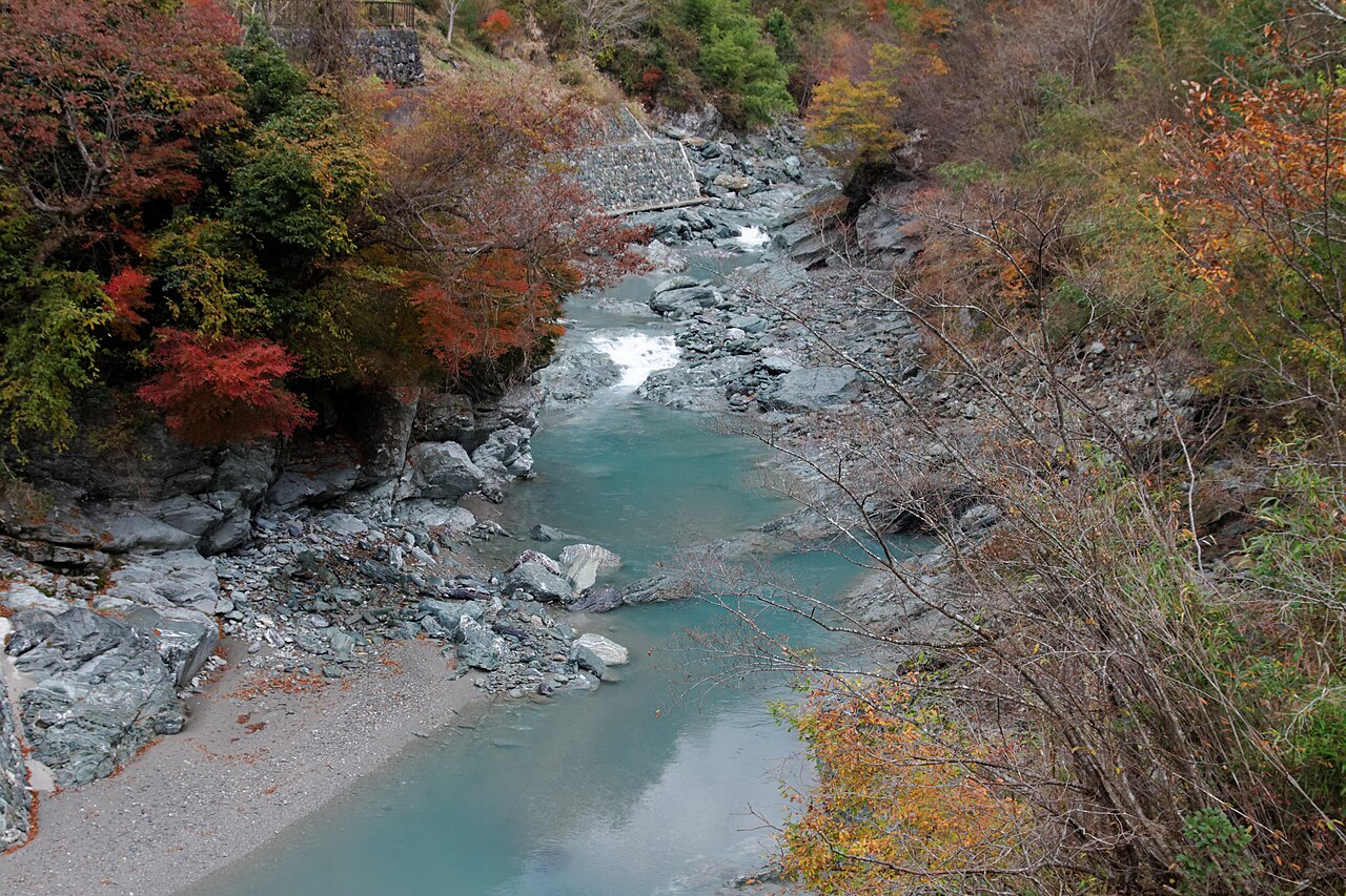 The Iya River running over grey rocks through the upper Iya Valley, with red and orange autumn leaves on the banks