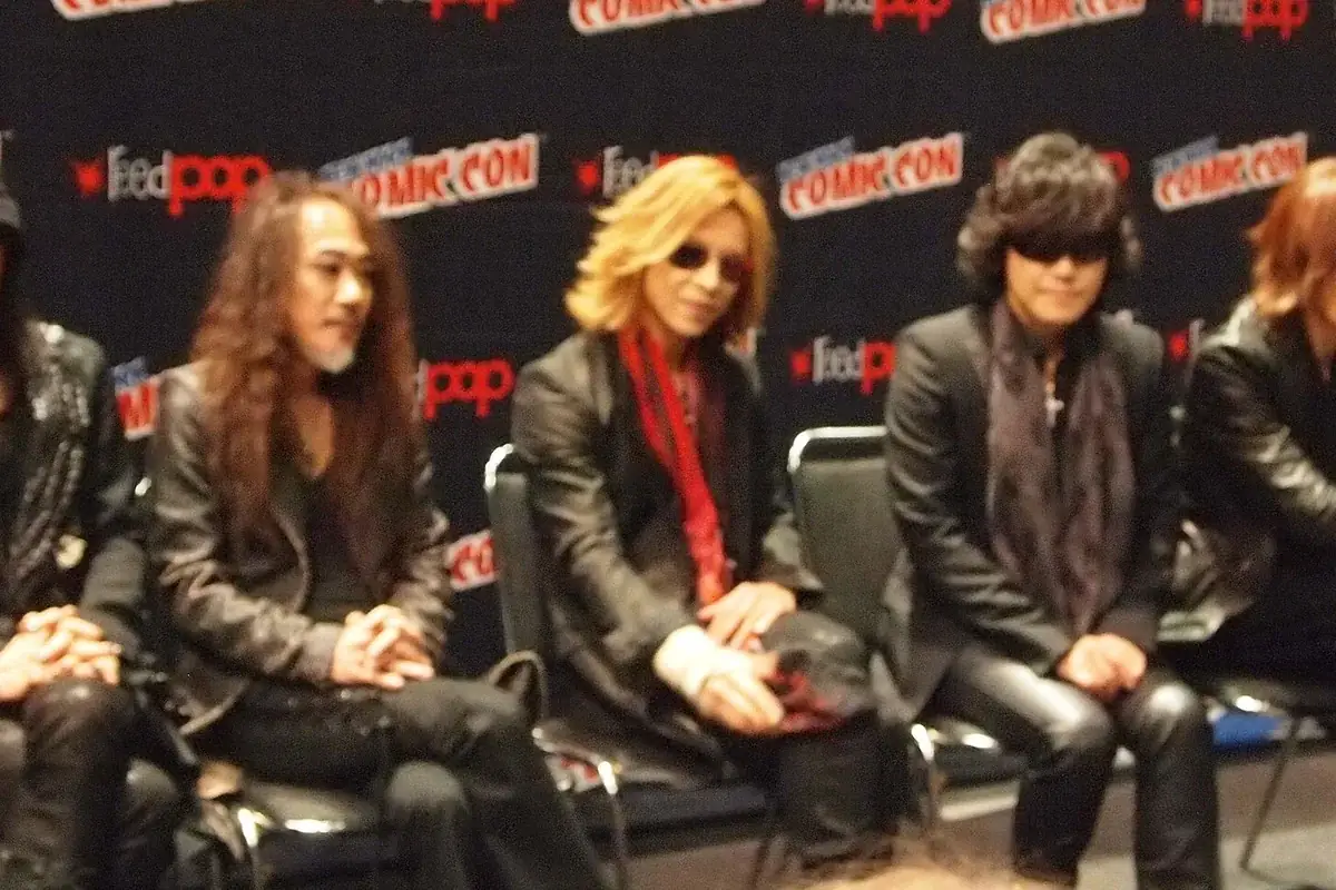 Members of X Japan seated in a row on a stage panel in dark clothes and sunglasses, in front of a New York Comic Con backdrop