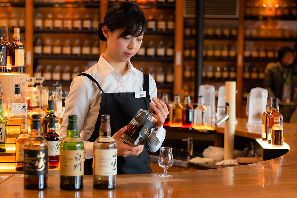 Bartender pouring a sample at the tasting bar of the Yamazaki Whisky Museum, with bottles of Hakushu and Yamazaki on the counter