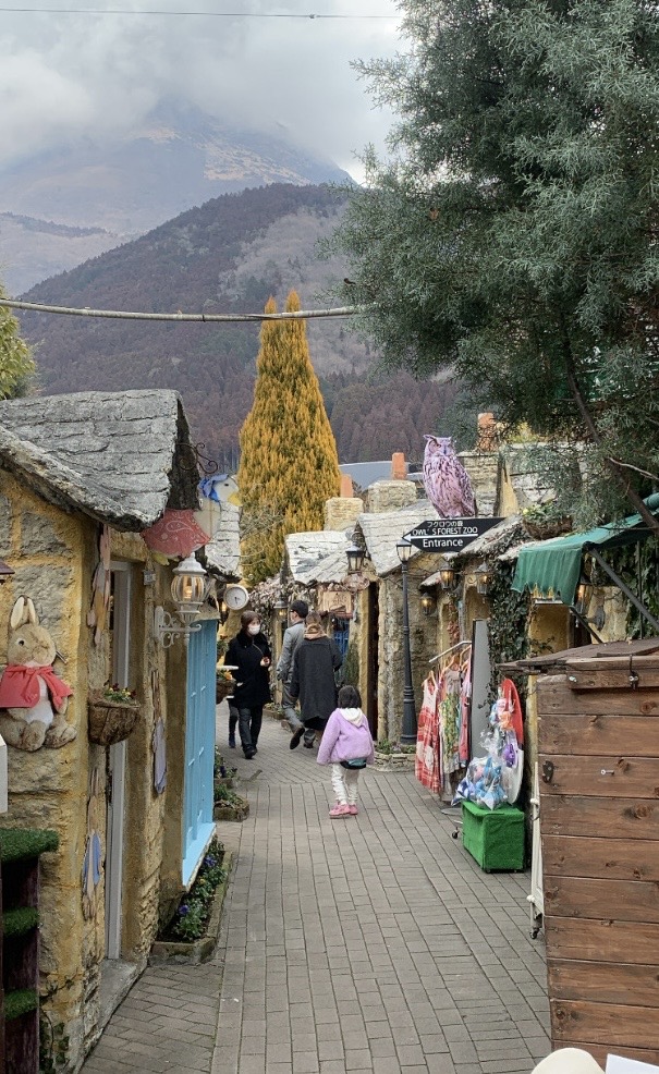 A paved lane in Yufuin Floral Village lined with Cotswolds style stone cottages and small shops, with mountains behind