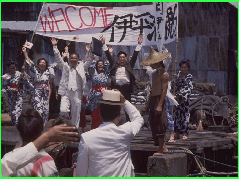 Colour still from Zegen (1987): a cheering crowd waving Japanese flags on a quayside under a Welcome banner